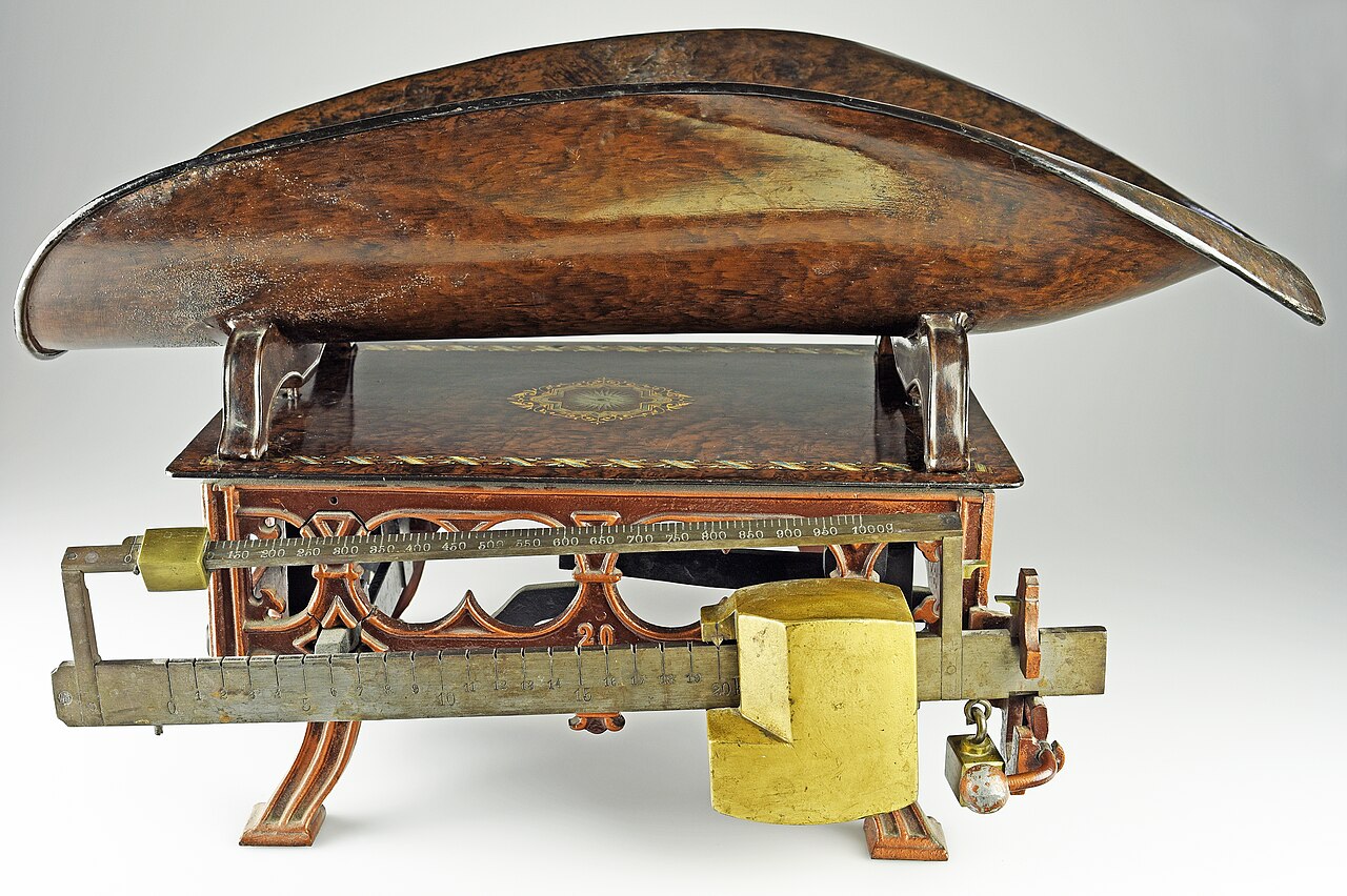 An antique infant weighing scale: a long, shallow tray of polished dark wood on a decorated base, with brass sliding weights on two graduated beams