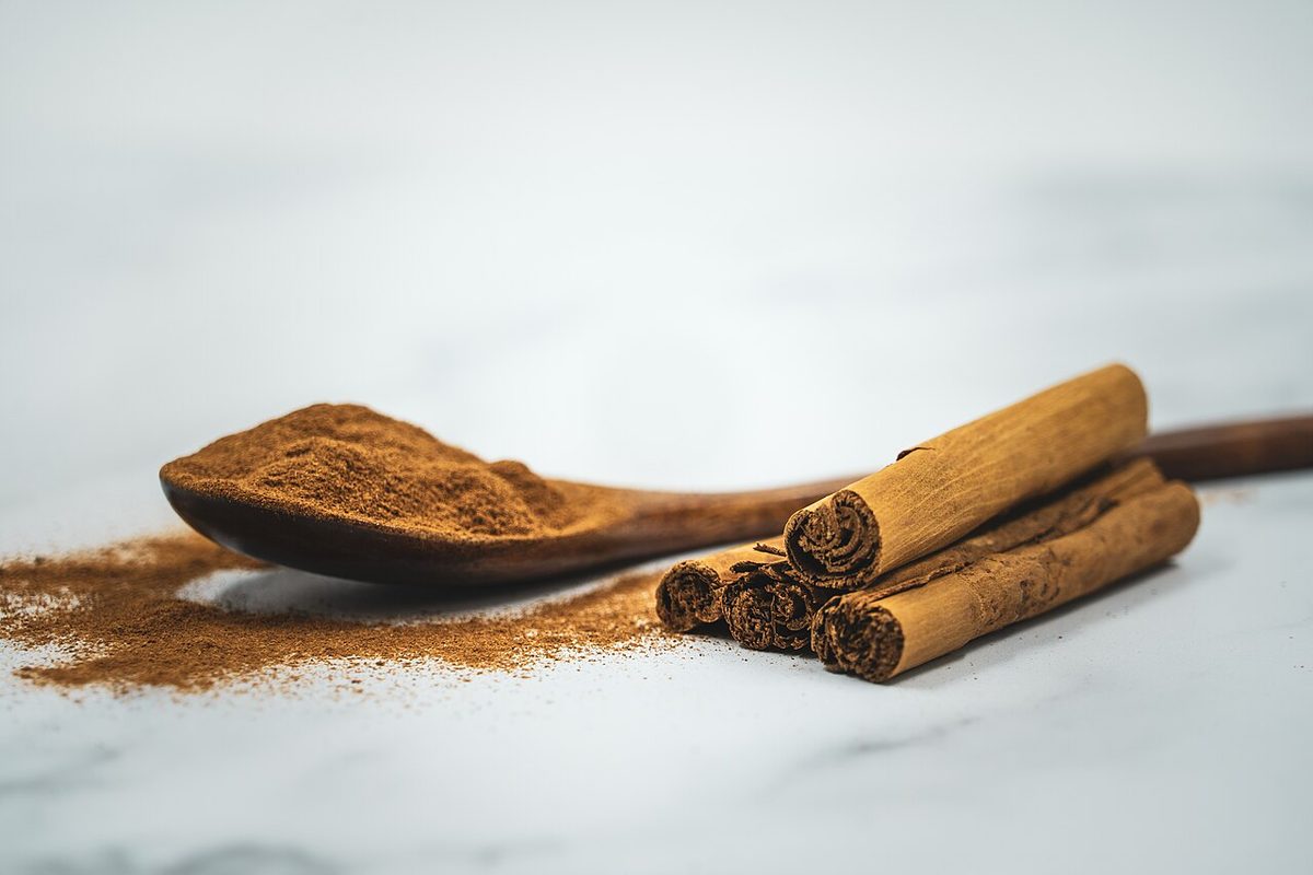 A wooden spoon heaped with ground cinnamon beside a small stack of cinnamon sticks on a white marble surface