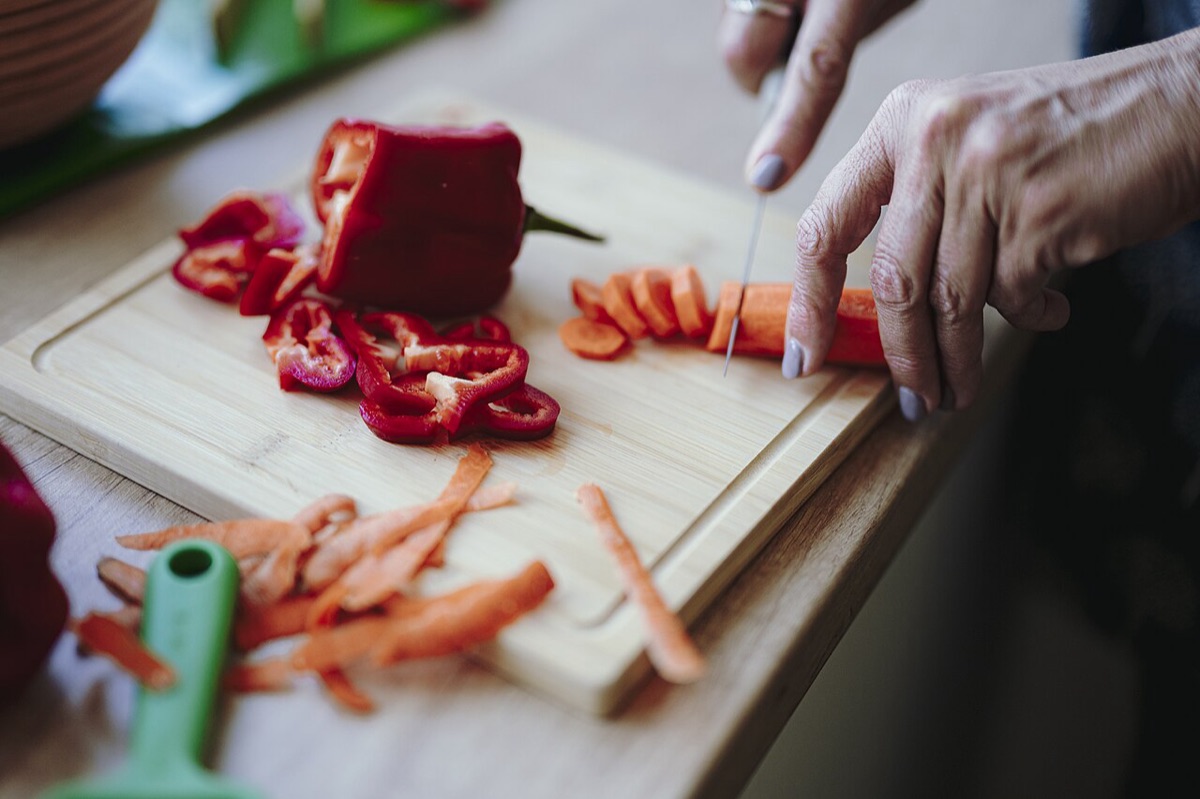 An older person's hands slicing a carrot on a wooden chopping board, with sliced red pepper and carrot peelings beside it