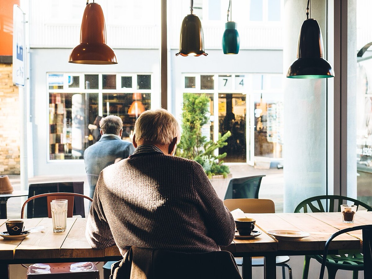 An older man with white hair, seen from behind, sits alone at a long wooden café table by a large window, with a cup of coffee and an empty plate in front of him