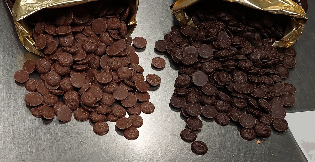 Two heaps of round chocolate drops spilling from gold-lined bags onto a steel worktop, milk chocolate on the left and darker chocolate on the right