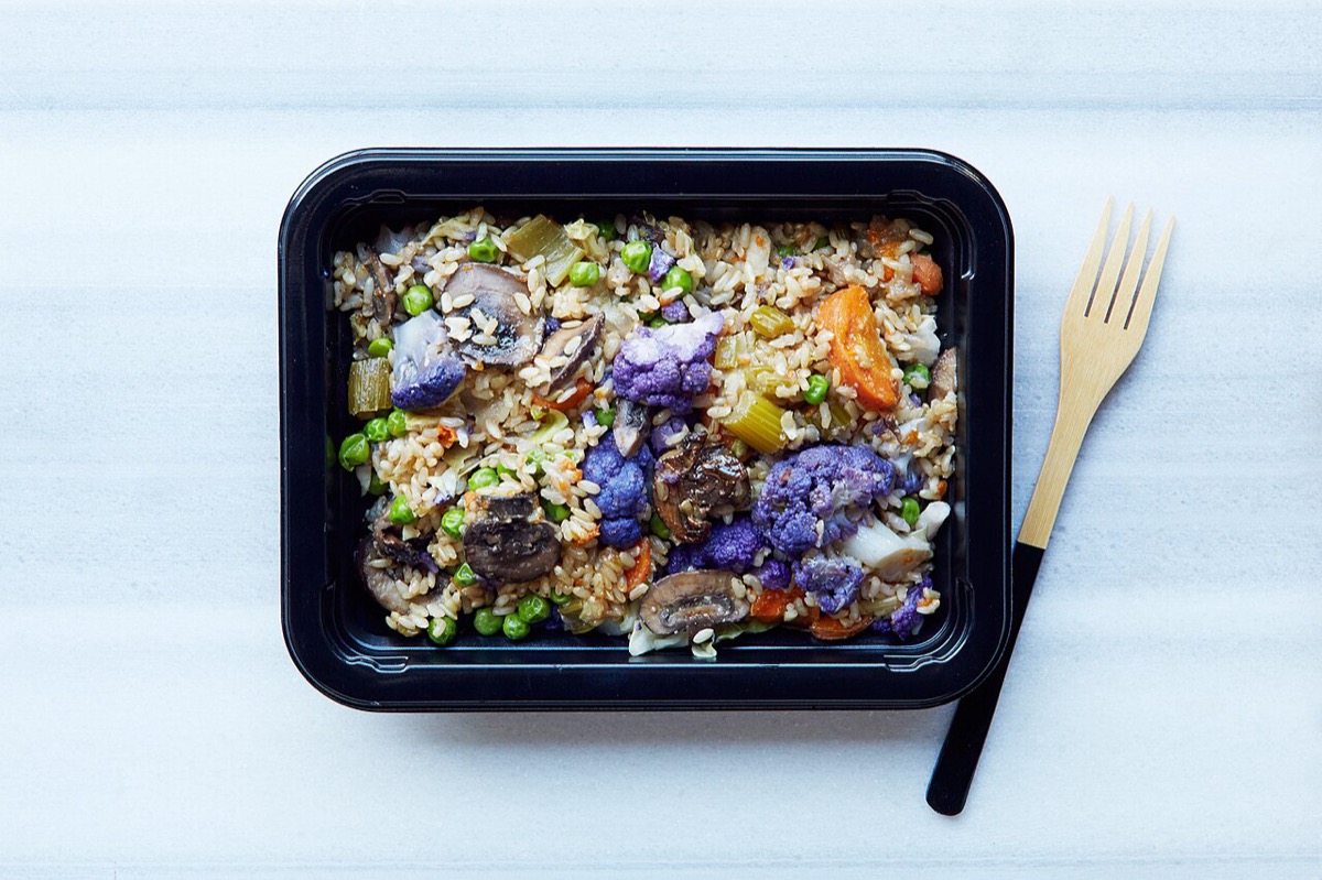 A ready meal of brown rice, purple cauliflower, mushrooms, peas and carrot in a black takeaway tray, with a wooden fork beside it