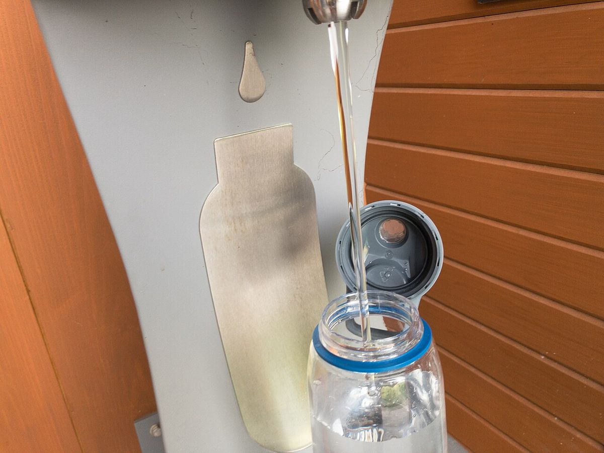 A clear reusable water bottle being filled from a stream of water at an outdoor bottle refill station set into a wooden wall