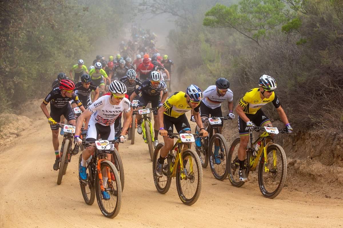 A pack of mountain bikers climbing a dusty dirt road between bushes during the Cape Epic