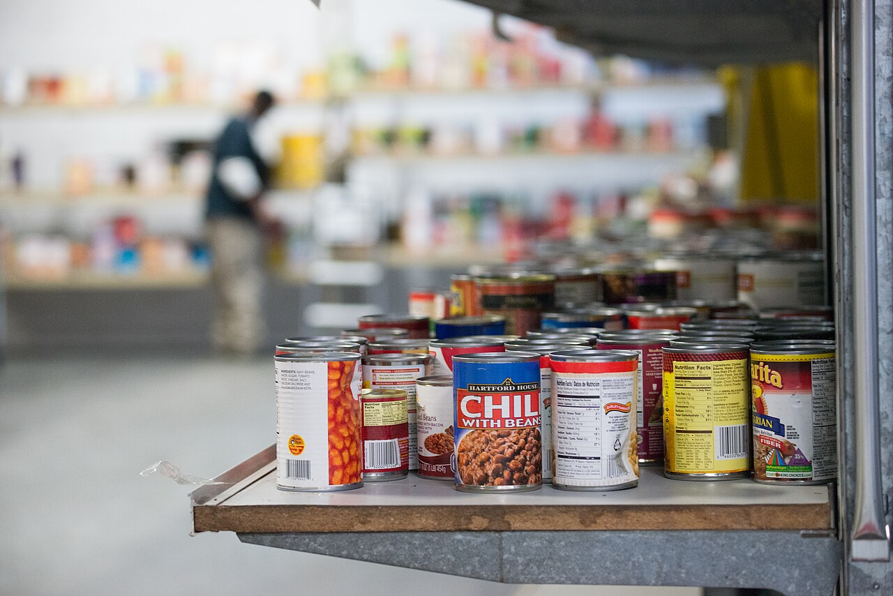 Tins of beans and chilli lined up on a metal shelf in a food bank warehouse, with a worker stocking shelves out of focus behind