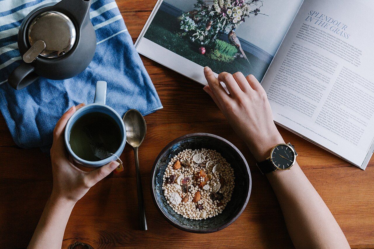 Seen from above, a bowl of puffed-grain cereal with nuts and dried fruit on a wooden table, with a cup of tea, a teapot and an open magazine