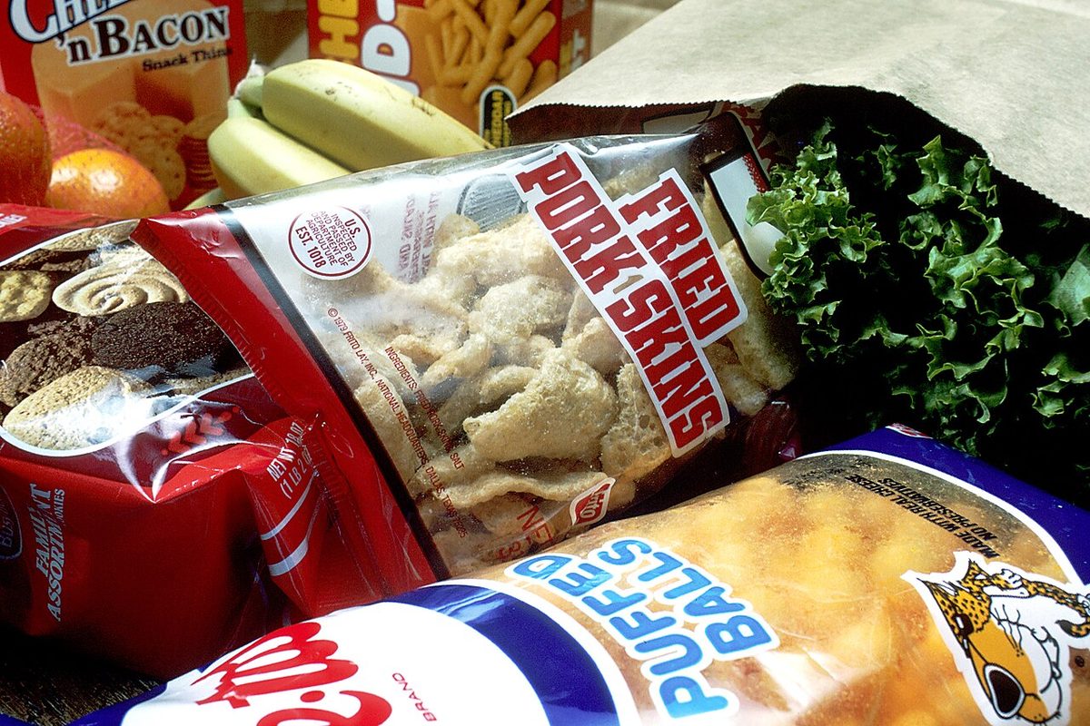 A paper grocery bag tipped over with packets of cookies, fried pork skins, cheese puffs and bacon crackers spilling out, with bananas, oranges and lettuce behind them