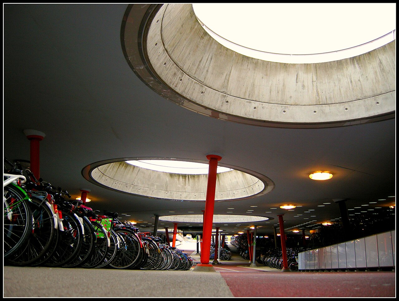 Rows of parked bicycles under round concrete skylights and red pillars in the bicycle parking at Groningen railway station