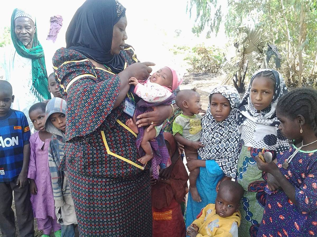 A health worker in a patterned dress and black headscarf holds a baby and gives oral vaccine drops, surrounded by children and an older man in a farming community in Adamawa, northern Nigeria