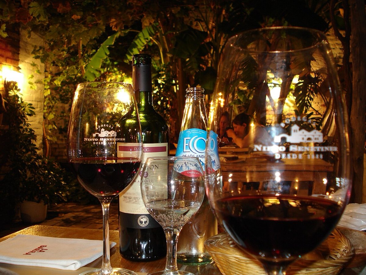 Two glasses of red wine and a small glass of water on a restaurant table at night, with a wine bottle and a bottle of mineral water behind them