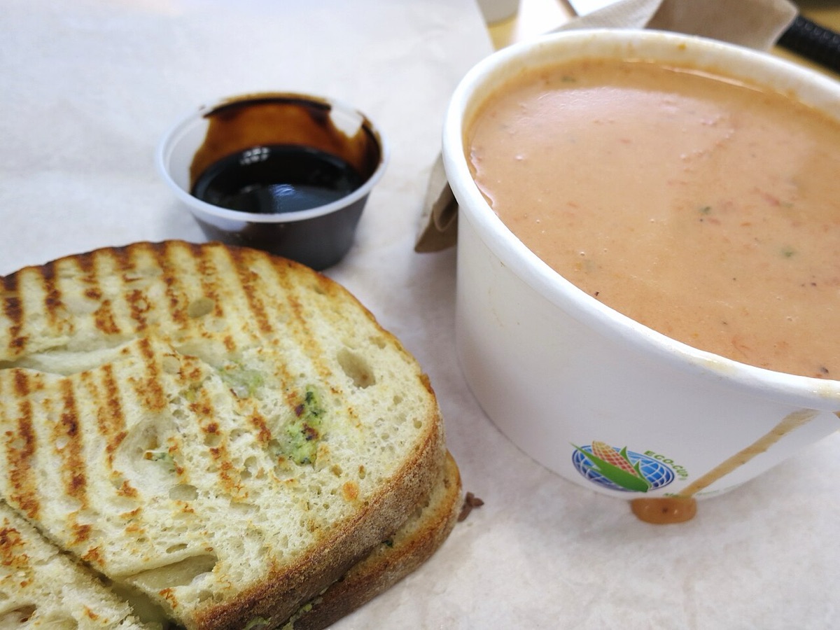 A toasted sandwich and a paper cup of tomato soup with a small pot of dark dip, eaten on a desk
