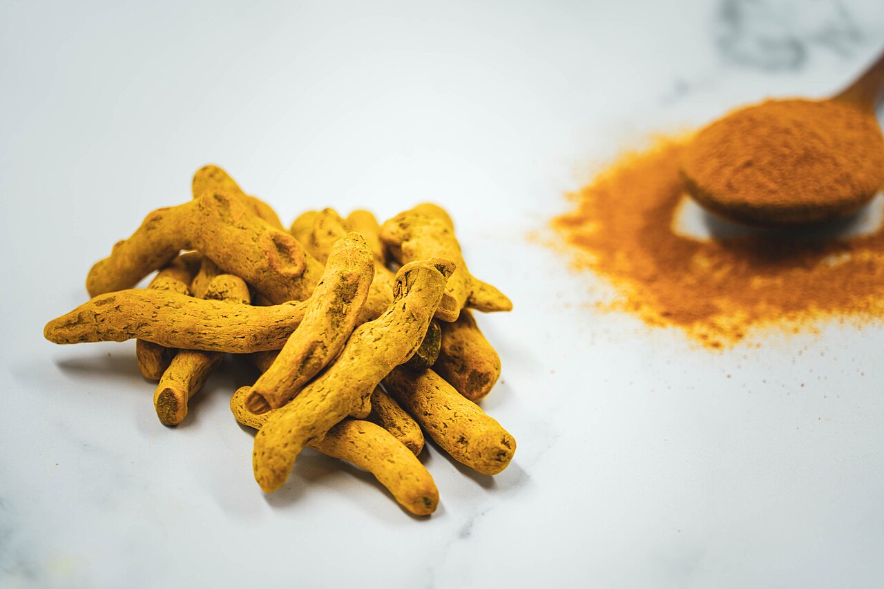 A small pile of dried turmeric roots, knobbly and mustard yellow, on a white marble surface, with a wooden spoon of ground turmeric spilling orange powder behind them