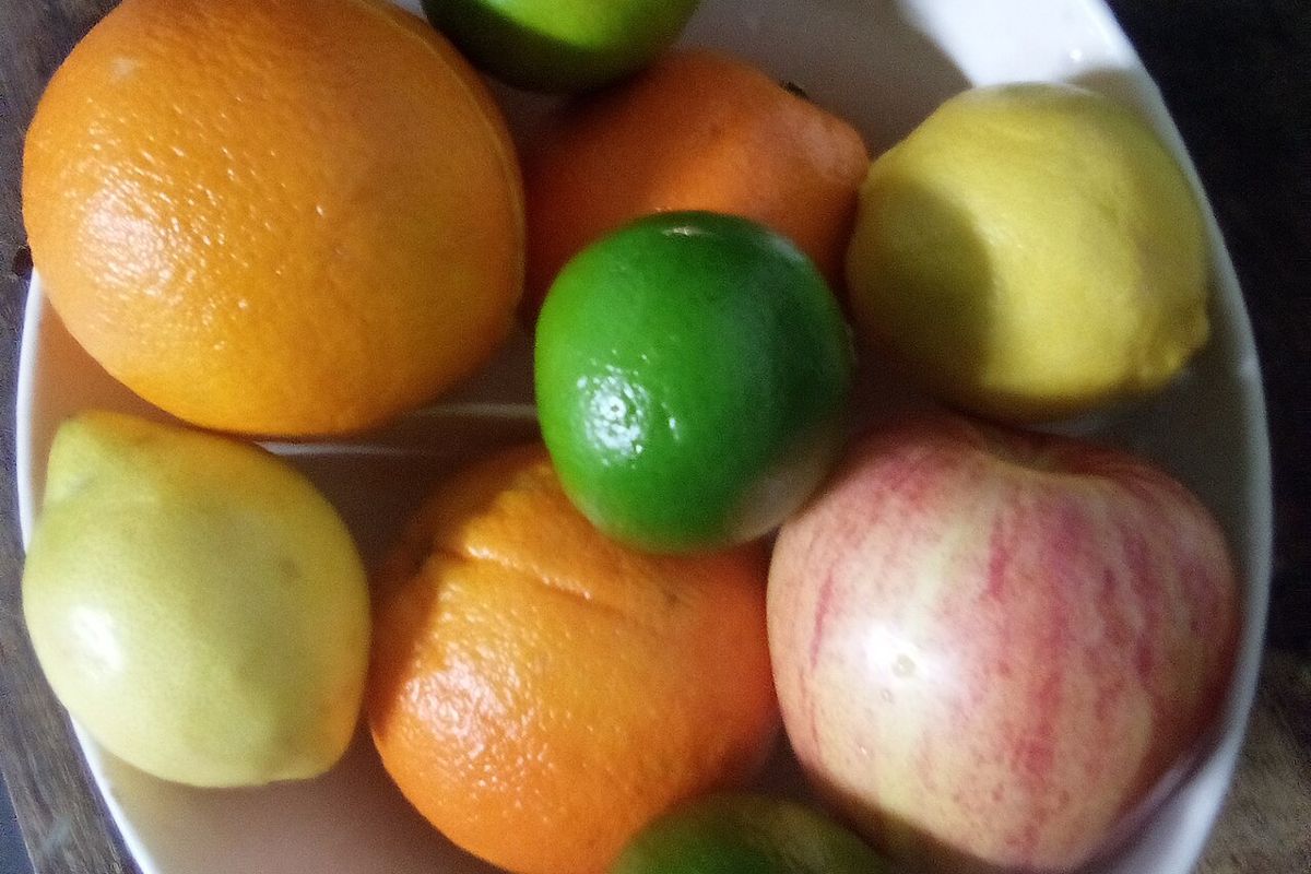 A white bowl of oranges, limes, lemons and an apple on a wooden table