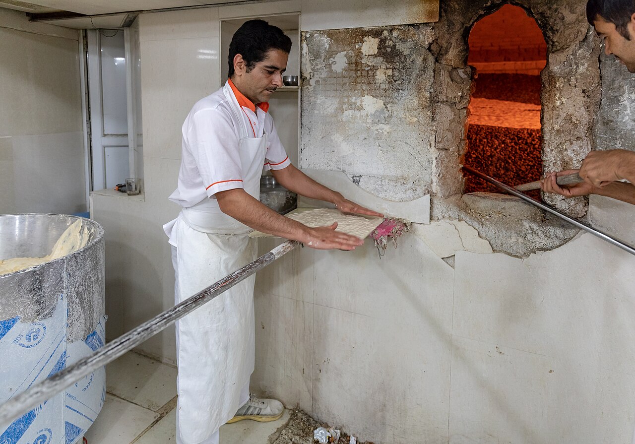 A baker in a white apron stretches flatbread dough on a paddle beside the glowing mouth of a pebble oven in a Tehran bakery