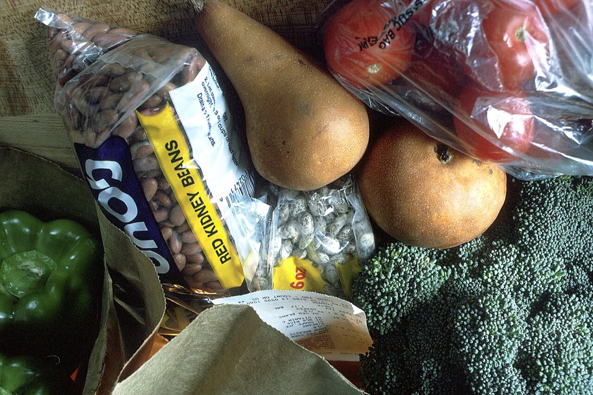 An open paper grocery bag spilling out red kidney beans, a bag of dried beans, pears, tomatoes, broccoli and green peppers onto a wooden table