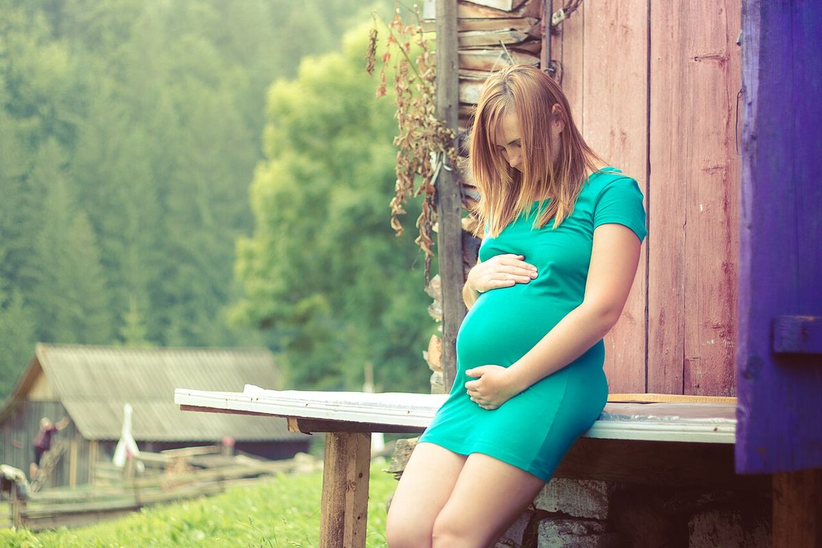 A pregnant woman in a green dress sits on a wooden bench outside a farmhouse, both hands resting on her belly