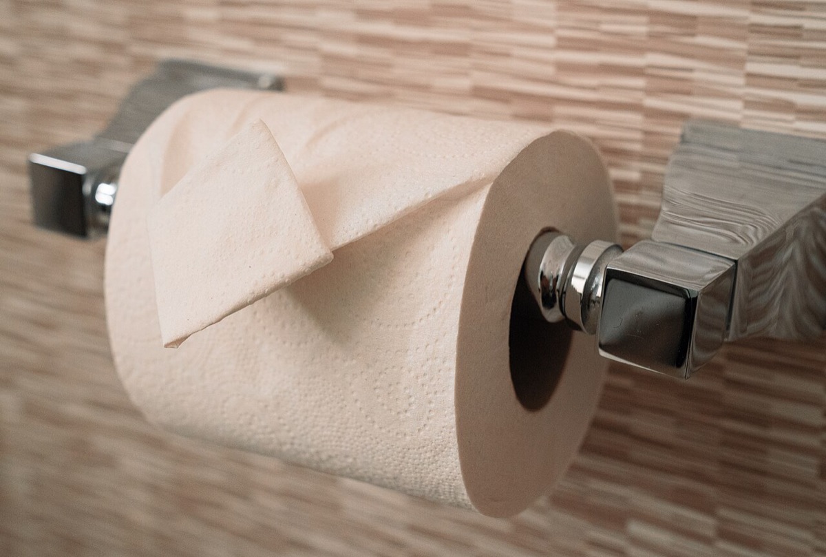 A roll of toilet paper on a chrome holder, its end sheet folded into a point, against a beige tiled wall