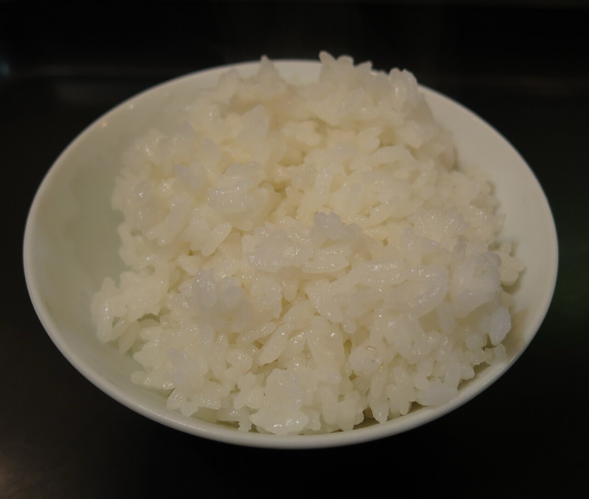 A white bowl heaped with cooked white rice on a dark table