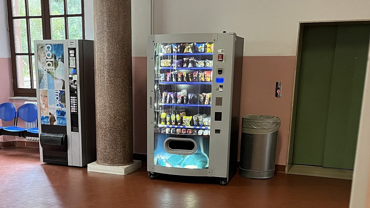 A coffee machine and a snack vending machine against a pink and white wall in the entrance hall of an Italian hospital