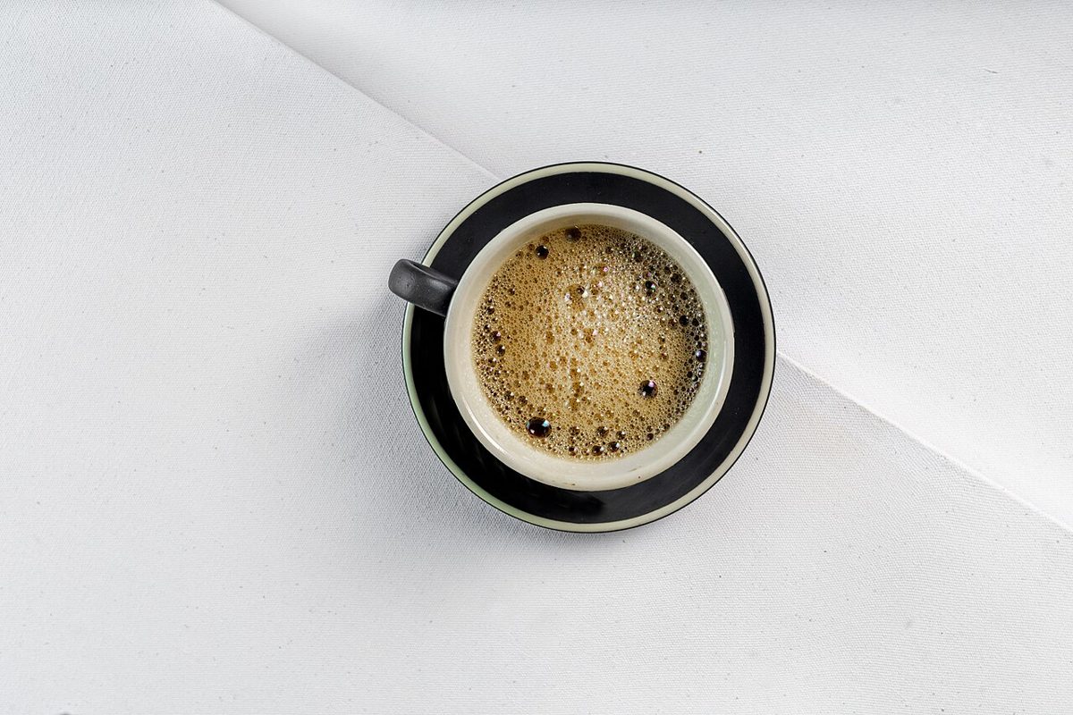 A cup of black coffee with a light froth on a dark saucer, seen from above on a white tablecloth