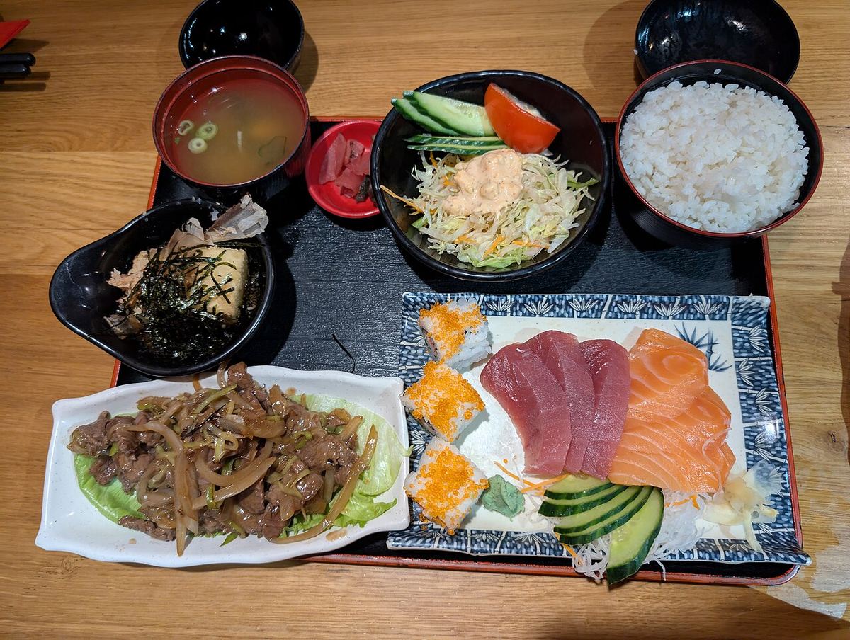 A Japanese set meal on a black tray seen from above: a bowl of rice, miso soup, a cabbage salad, tofu with seaweed, stir-fried beef with onions, and sliced raw tuna and salmon