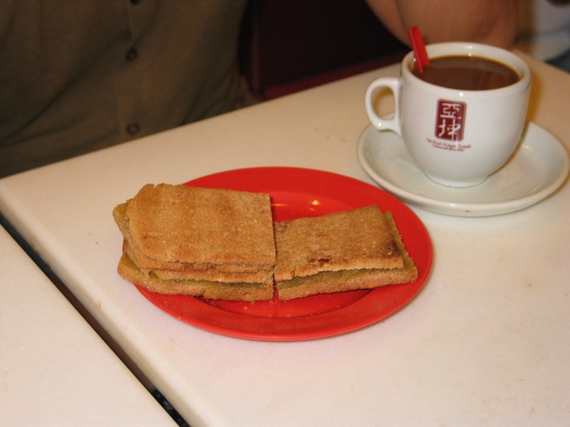 Two stacks of toasted bread with a green coconut jam filling on a red plastic plate, beside a white cup of coffee on a saucer, on a white café table