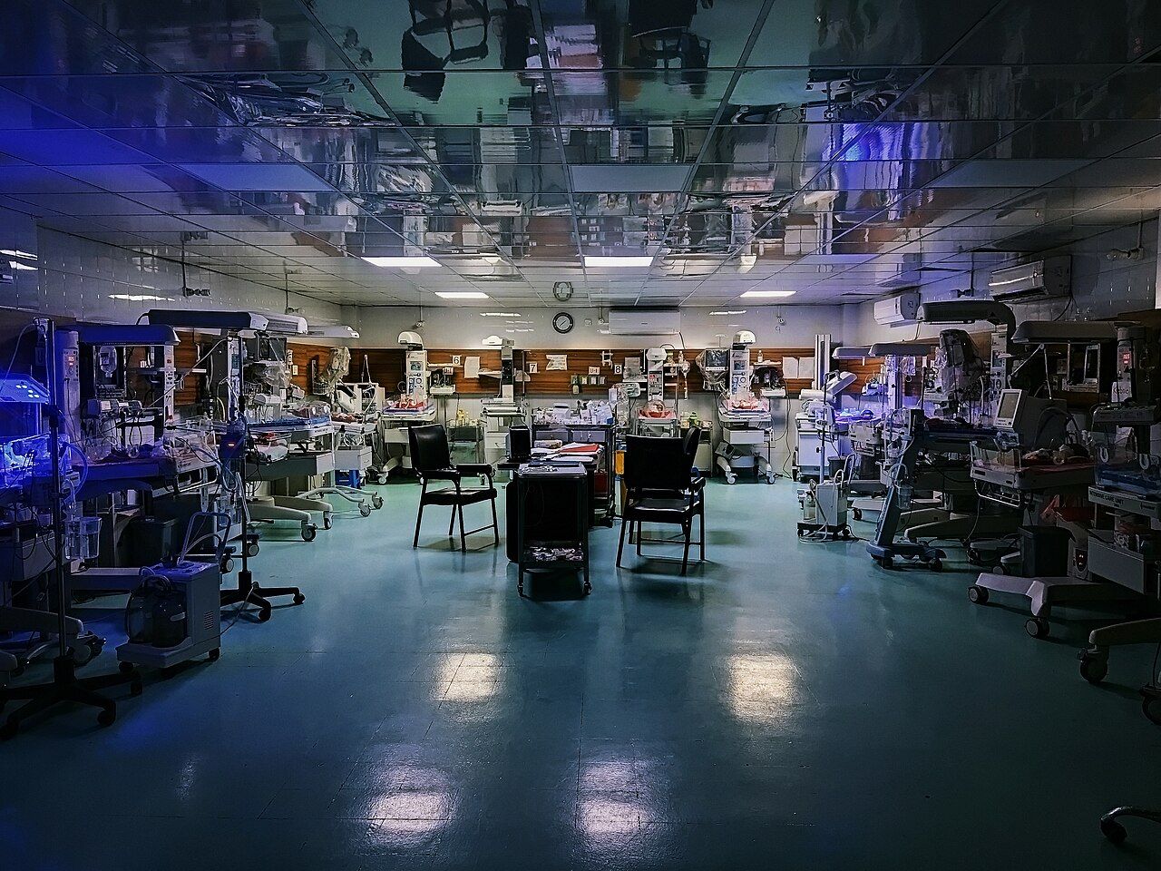 A dimly lit neonatal intensive care unit at night, with incubators and monitors along the walls and two empty chairs at a desk in the middle of the room