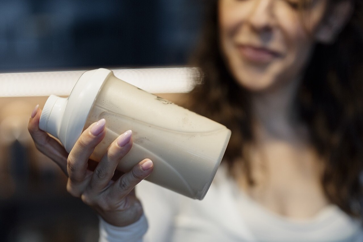 A woman, out of focus, smiles as she shakes a white shaker bottle full of a pale drink in a dim kitchen