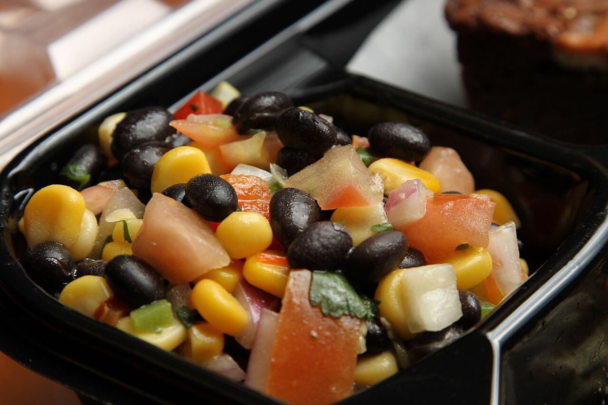 Close-up of a corn and black bean salad with chopped tomato, red onion and coriander leaves in a black takeaway tray