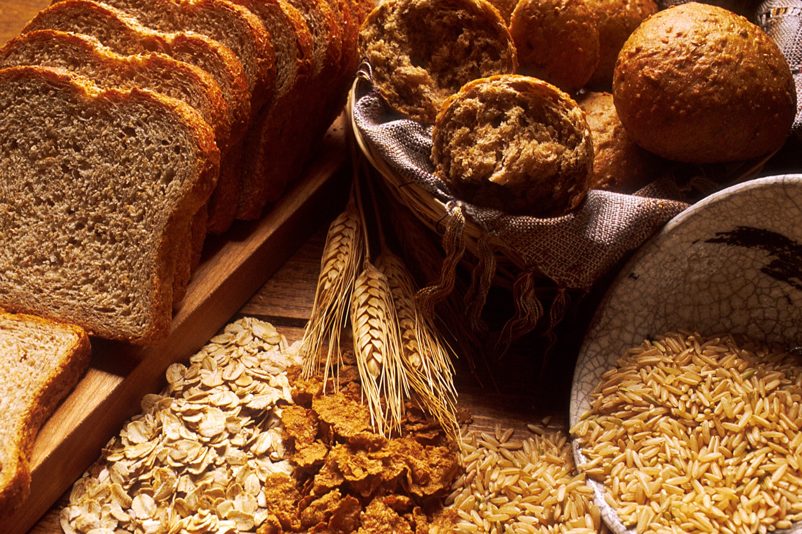 A sliced loaf of brown bread, a basket of rolls and four kinds of grain on a wooden table