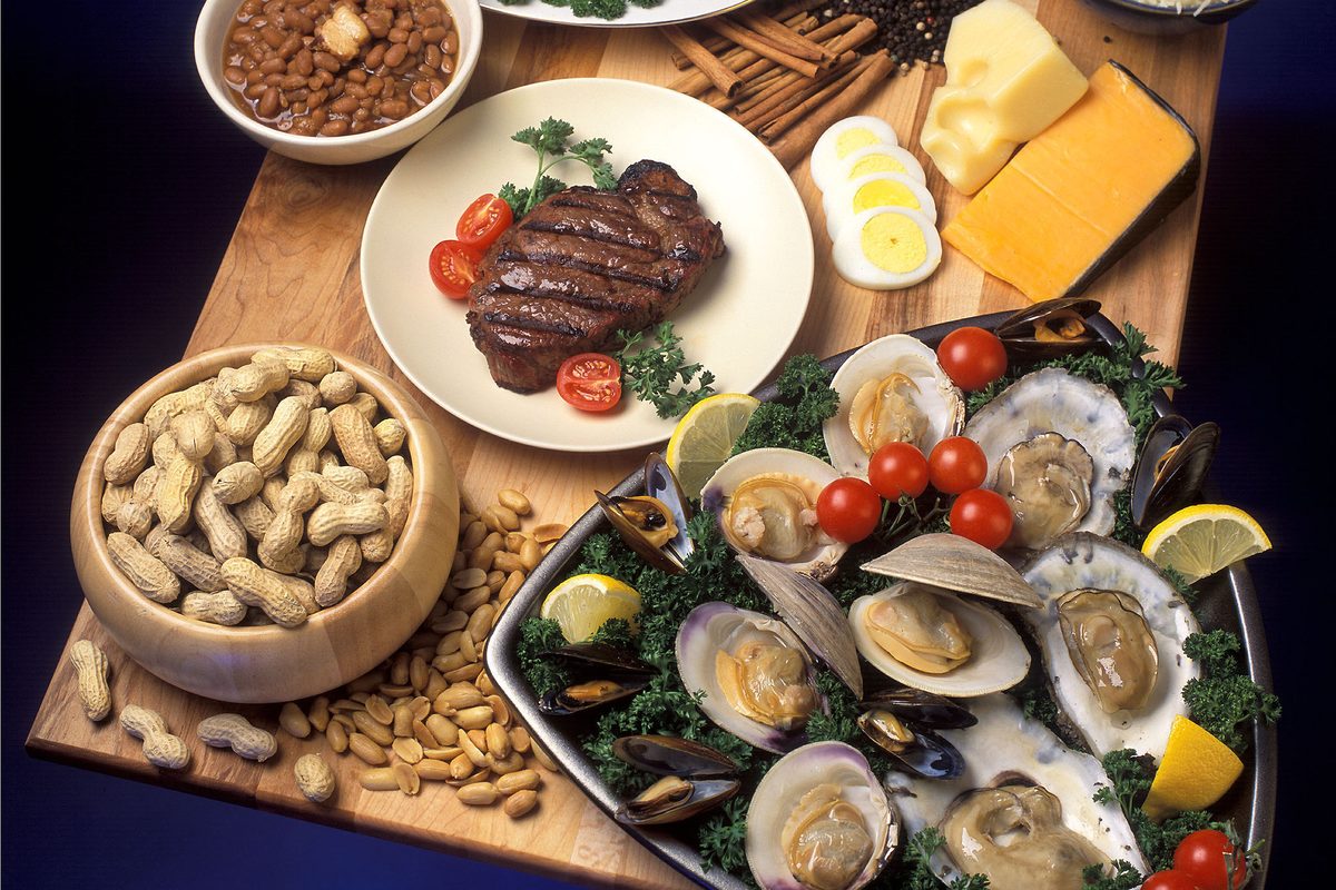 A wooden board laid with zinc-rich foods: a platter of oysters, clams and mussels, a grilled steak, a bowl of peanuts, cheese, boiled eggs and beans